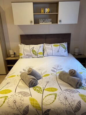 A neatly made bed with floral-patterned bedding, two pillows, and two rolled towels. Above the bed, there is a shelf with two decorative signs.