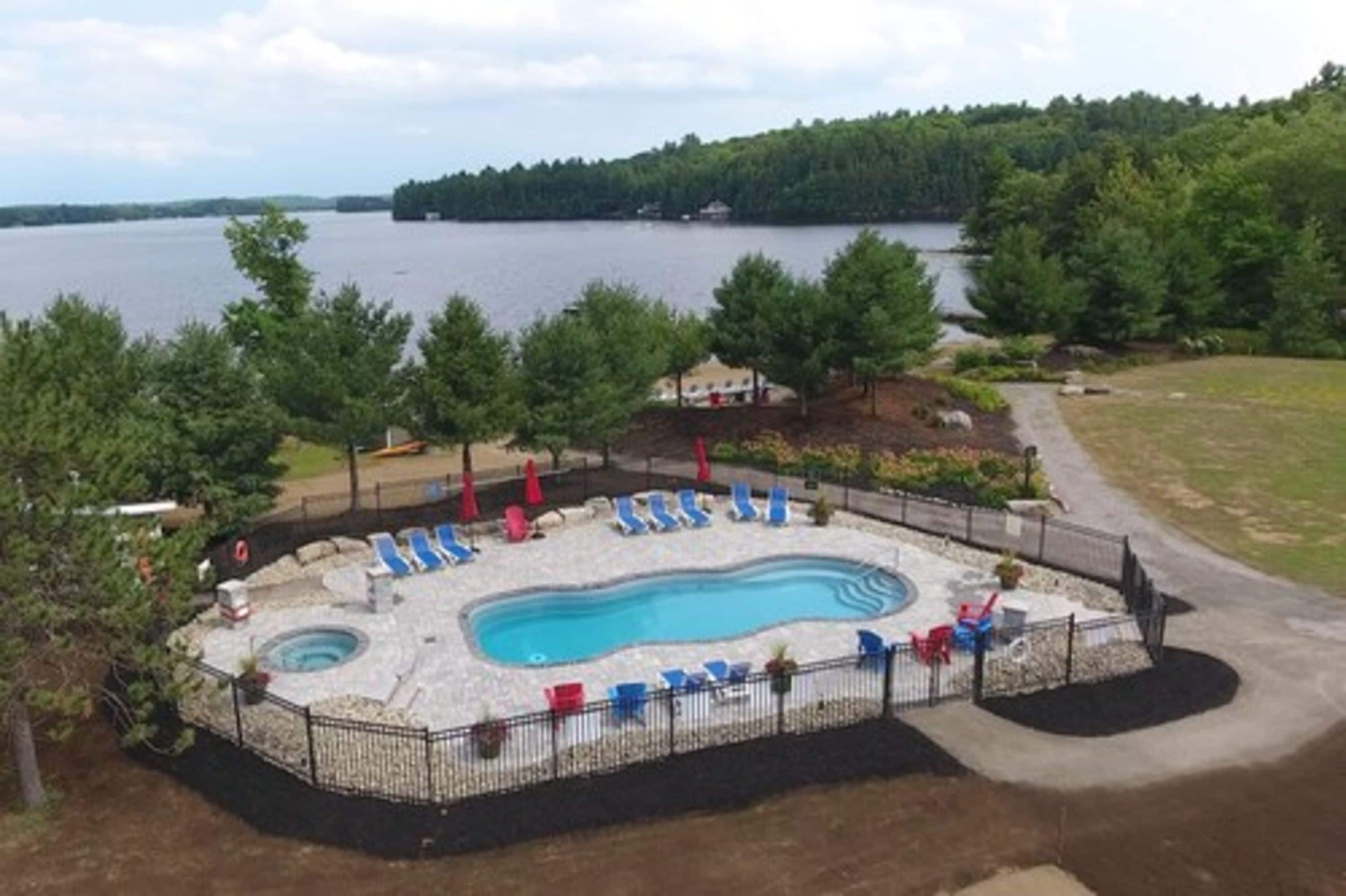 A swimming pool with lounge chairs, surrounded by a fence, with a view of a lake and forested area.