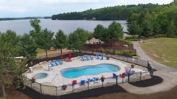 A swimming pool with lounge chairs, surrounded by a fence, with a view of a lake and forested area.