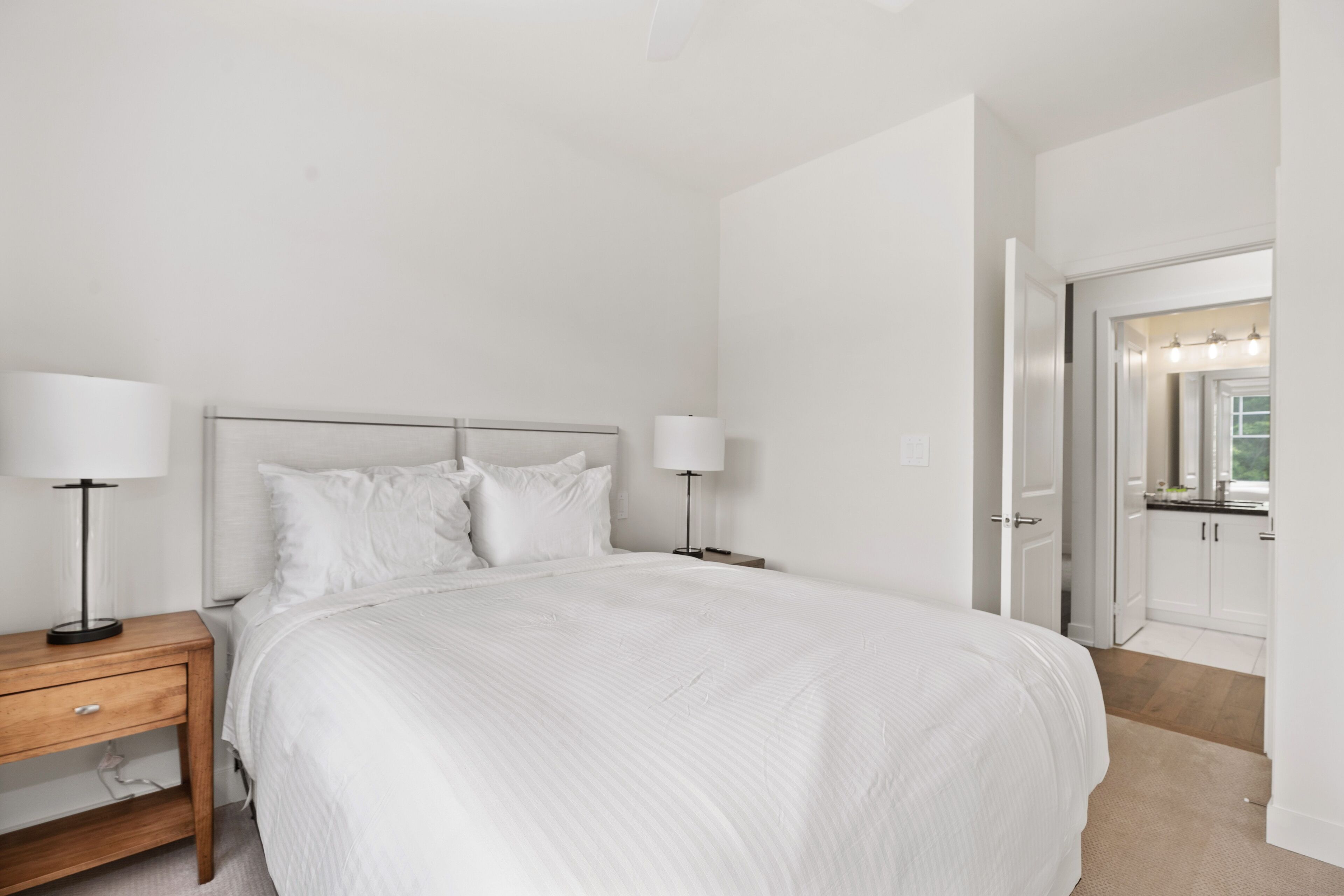 A large bed with white bedding, two bedside tables with lamps, a wooden nightstand, and a door leading to a bathroom with a mirror and lights.