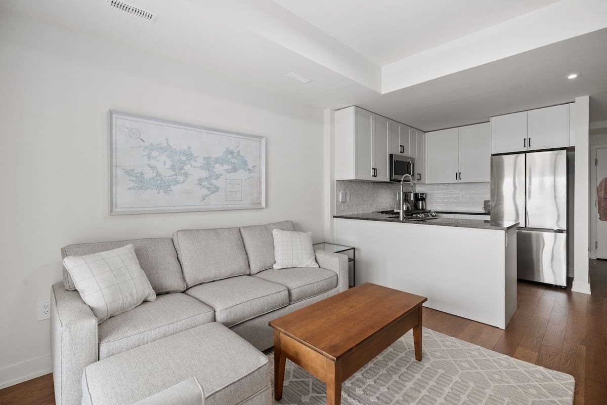 A modern living room with a grey sofa, a wooden coffee table, and a kitchen area with white cabinets and stainless steel appliances.