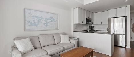 A modern living room with a grey sofa, a wooden coffee table, and a kitchen area with white cabinets and stainless steel appliances.