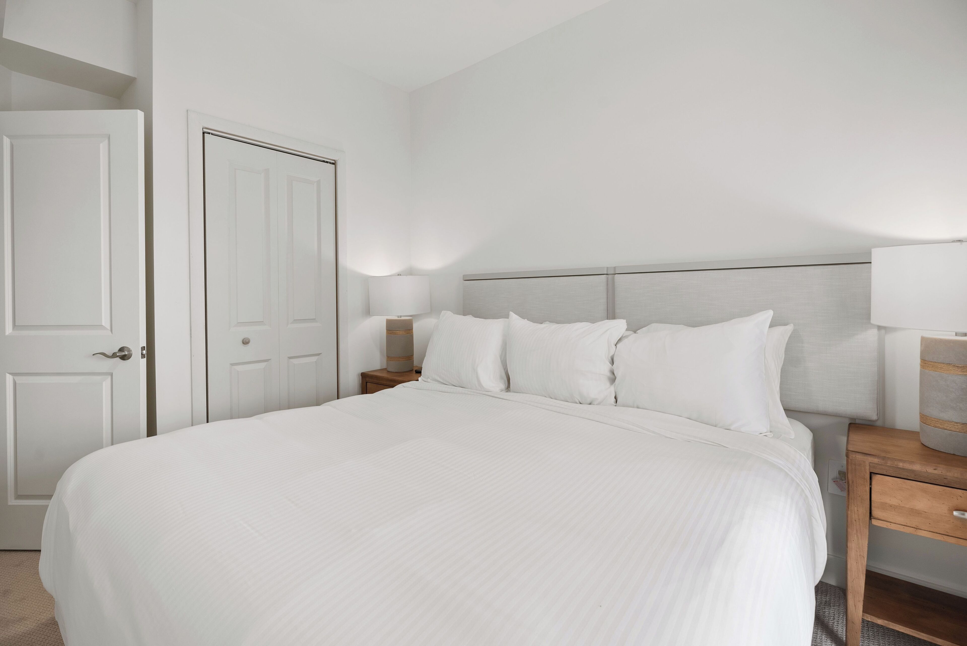 Una cama bien tendida con sábanas blancas, una mesita de noche de madera con una lámpara y una puerta a la izquierda.