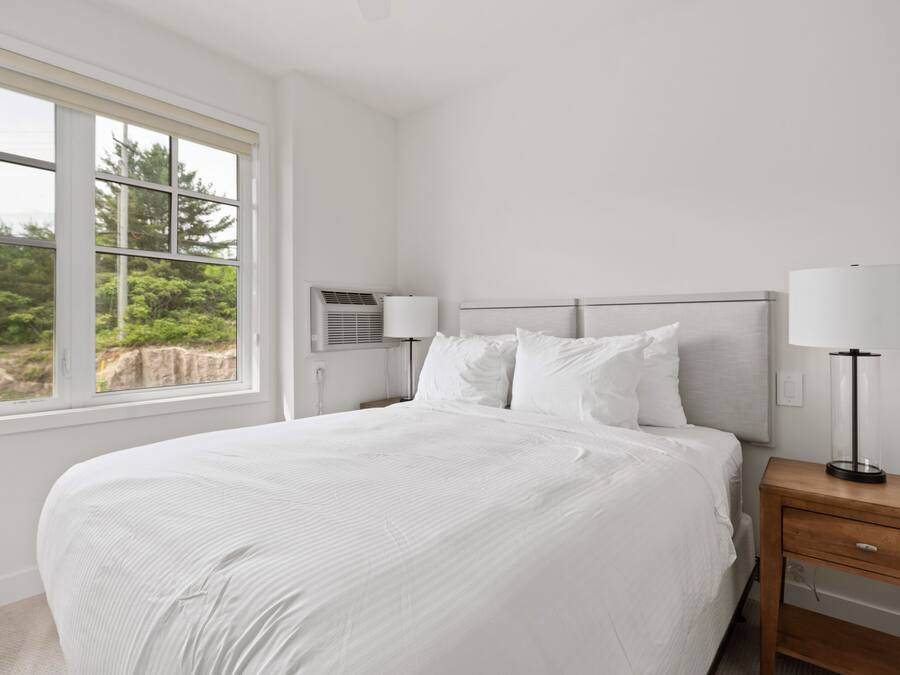 Un dormitorio con una cama grande, dos mesitas de noche con lámparas, una ventana con vistas a la vegetación y un equipo de aire acondicionado.