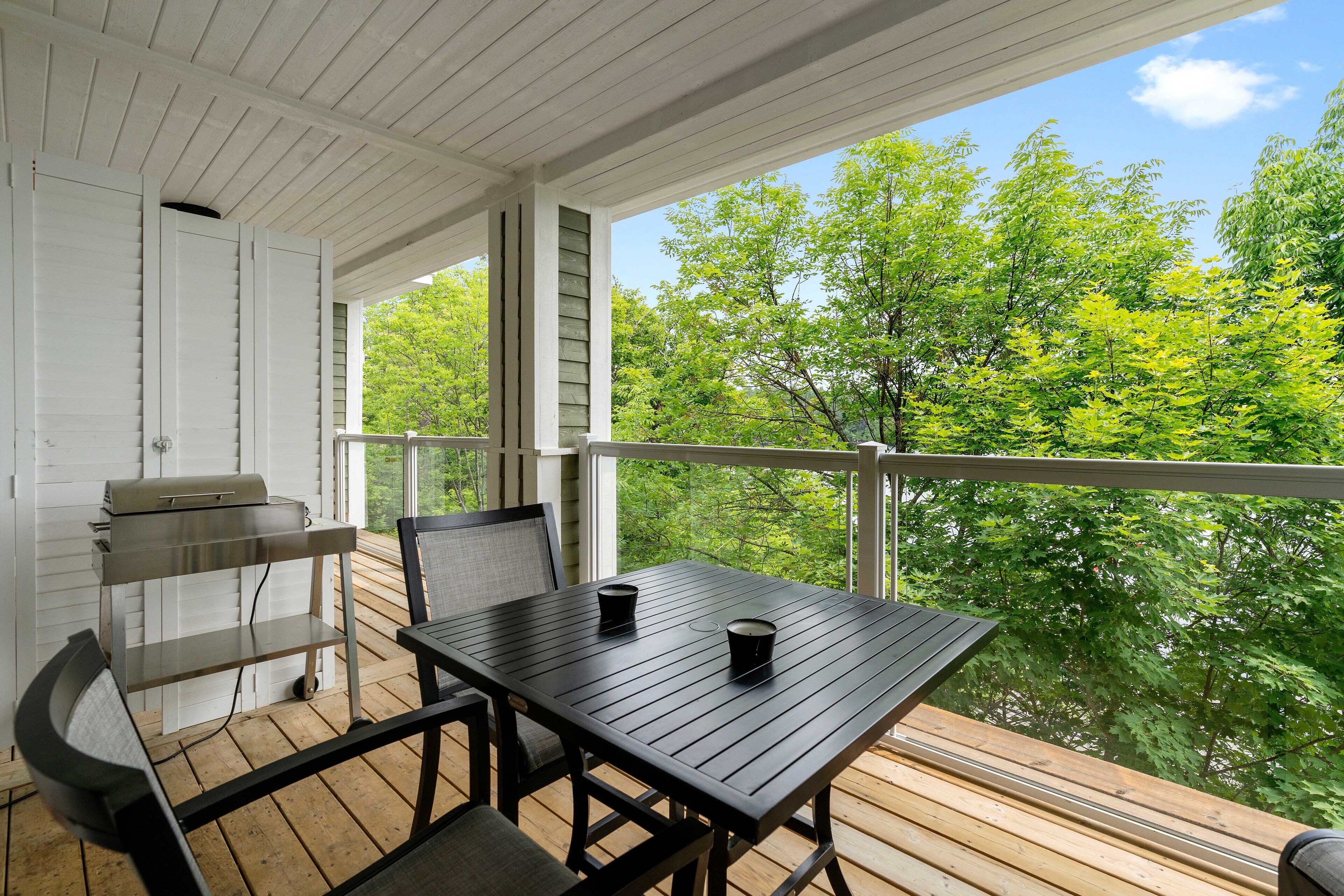 A wooden deck with a table and chairs, a grill, and a view of green trees.