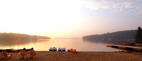 Una platja amb cadires i barques, un llac tranquil i una riba boscosa.