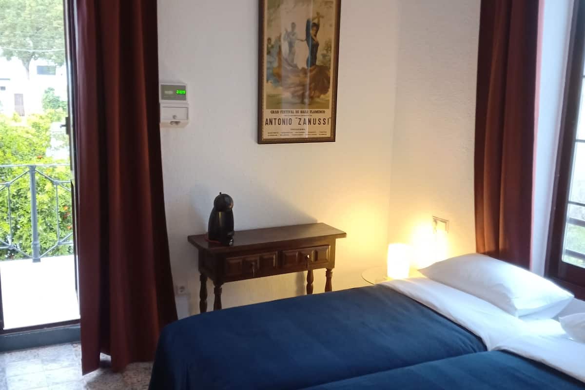 Hotellihuone, jossa on kaksi sänkyä, puinen yöpöytä, taulu seinällä ja näkymä vehreään maisemaan.