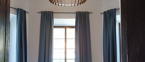 A bedroom with a wooden ceiling, a large window with curtains, a bed with white linens, and a hanging light fixture.