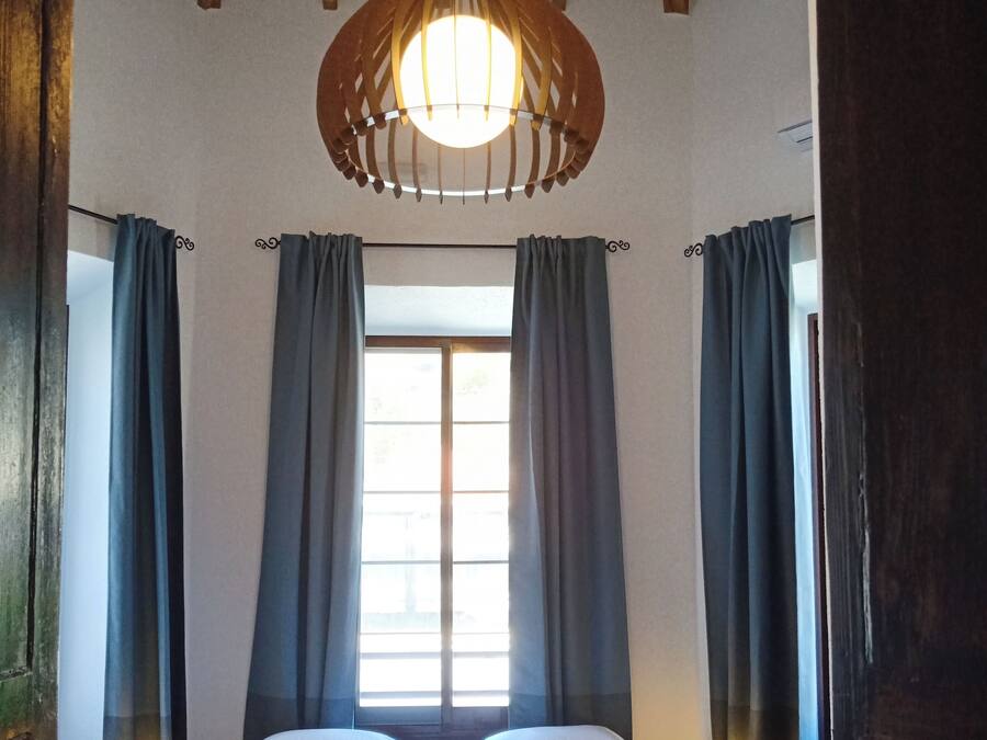 Un dormitorio con techo de madera, un ventanal con cortinas, una cama con sábanas blancas y una lámpara colgante.