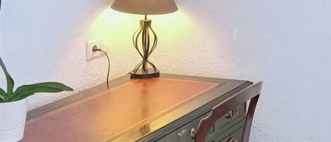 A wooden desk with a lamp, a chair, a potted plant, and a framed picture on the wall.