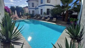 Una piscina con sillas de descanso, rodeada de plantas tropicales y un edificio de dos pisos con detalles azules.