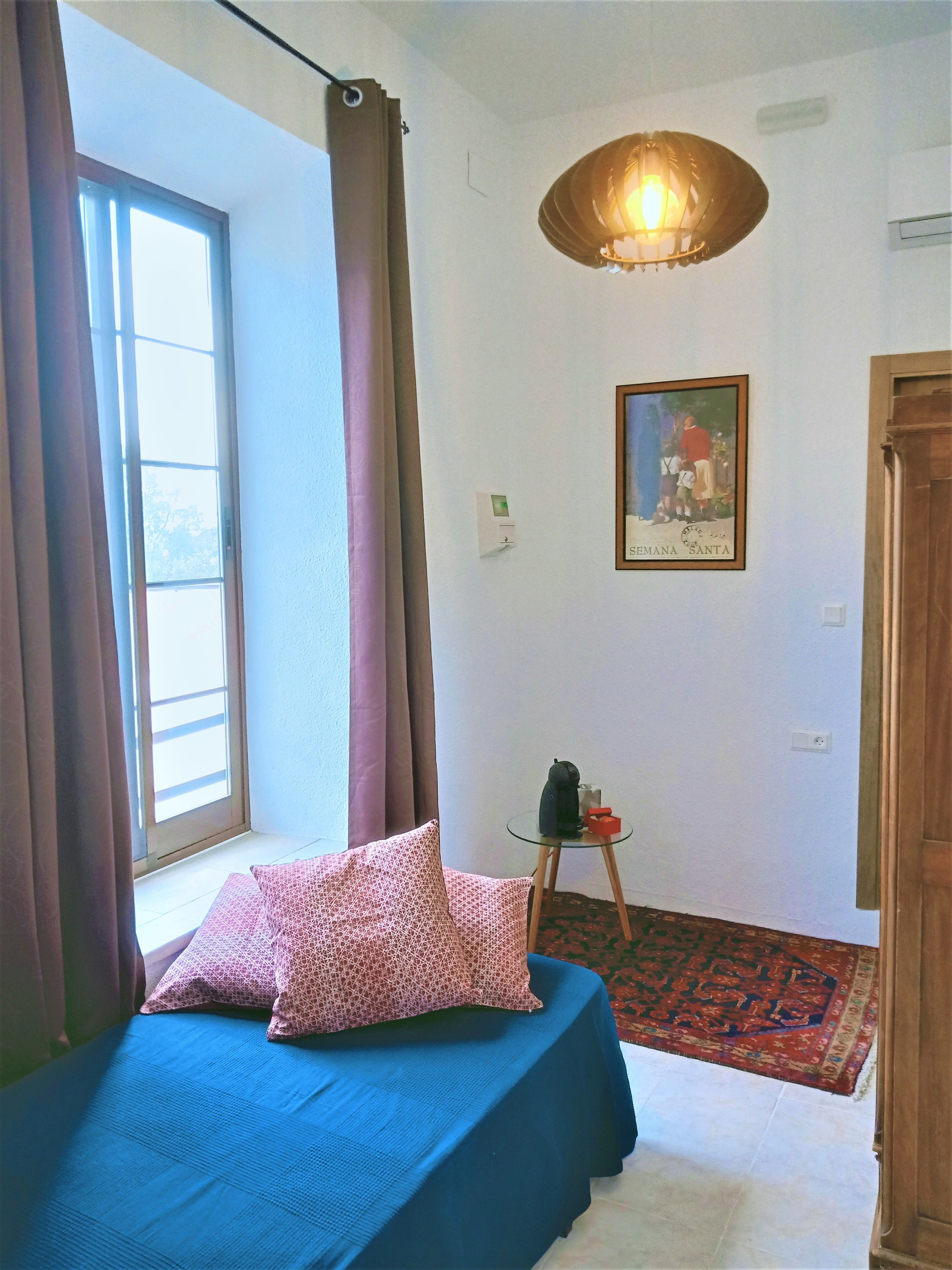 Un dormitorio con cama, ventana con cortinas, lámpara colgante, un cuadro en la pared y una mesita con hervidor y caja.
