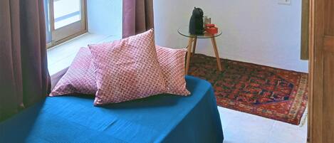Un dormitorio con cama, ventana con cortinas, lámpara colgante, un cuadro en la pared y una mesita con hervidor y caja.