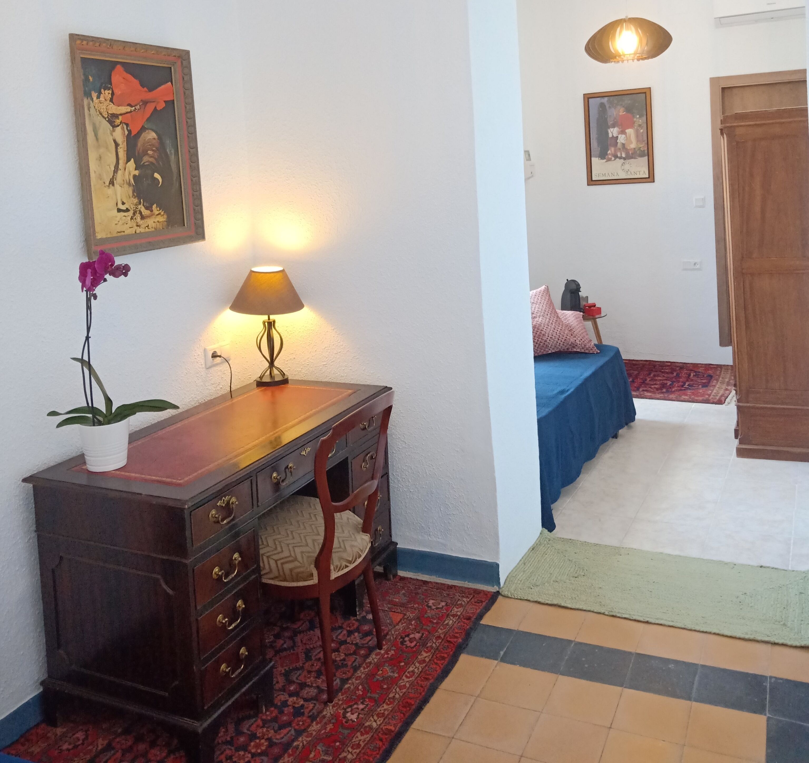 A room with a wooden desk, a lamp, a chair, a bed with a blue bedspread, a red rug, and a patterned rug.