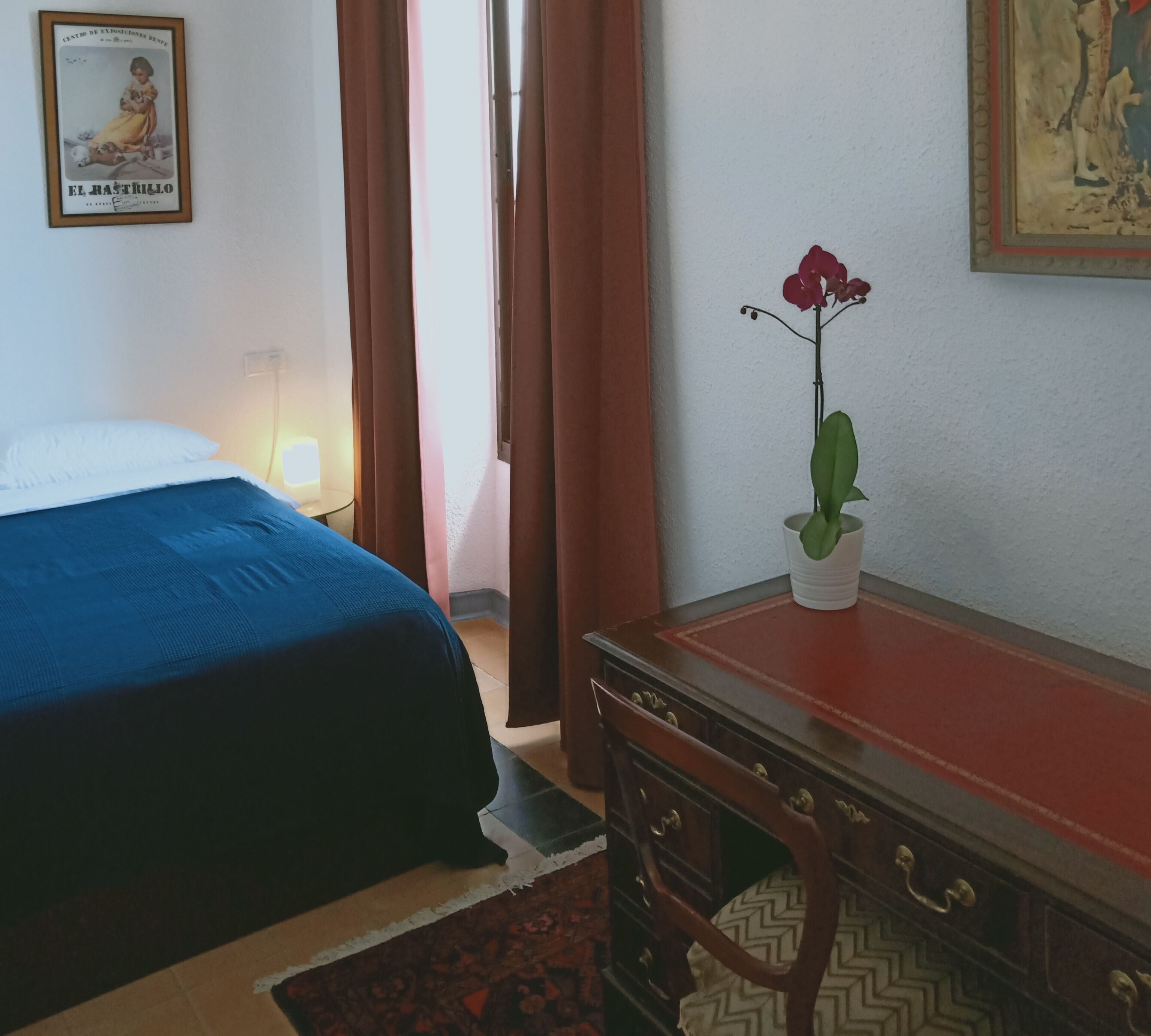 A hotel room with a bed, a dresser, a potted plant, and a framed picture on the wall.