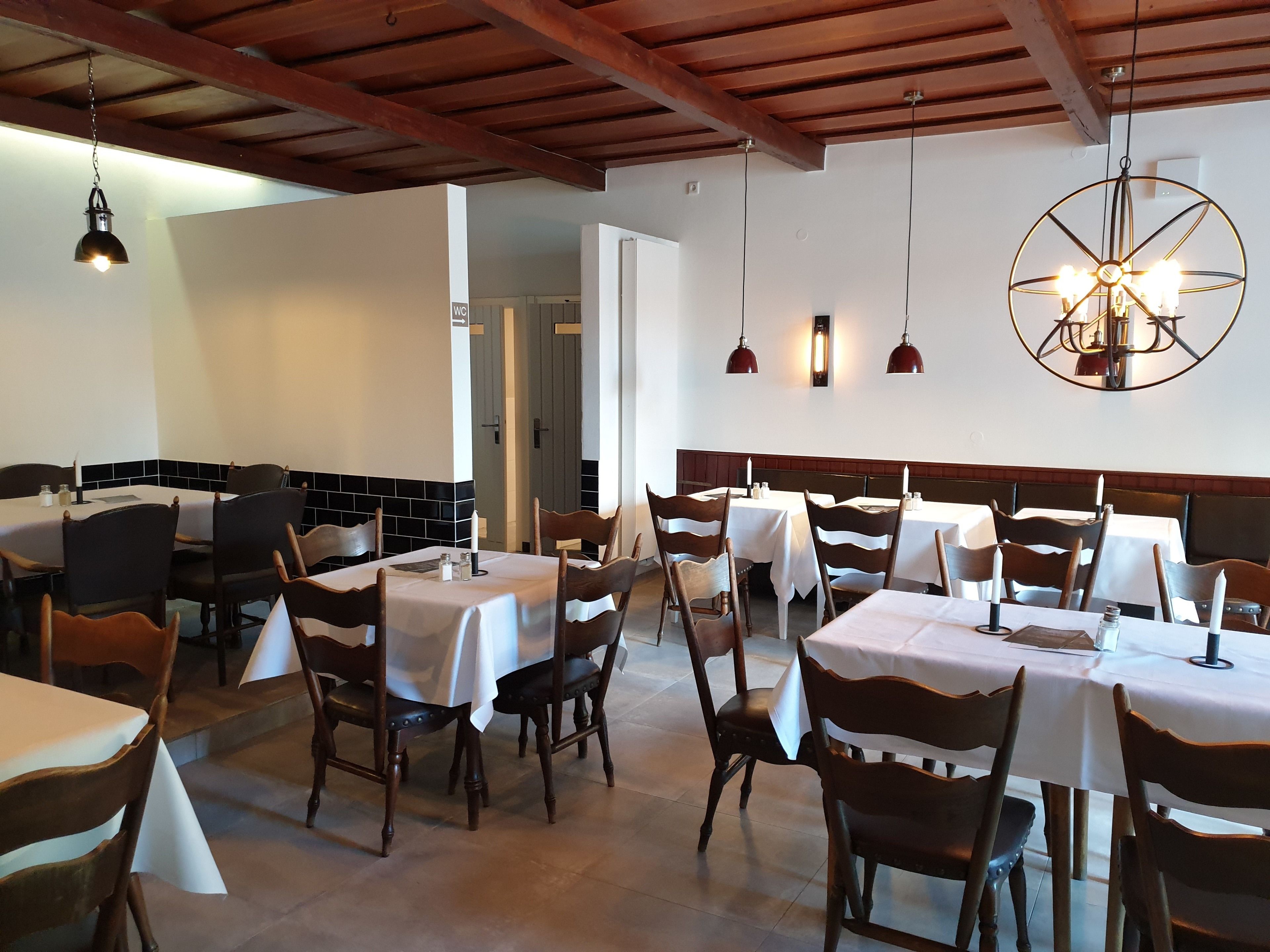A restaurant with multiple tables and chairs, wooden beams on the ceiling, pendant lights, and a wall-mounted light fixture.