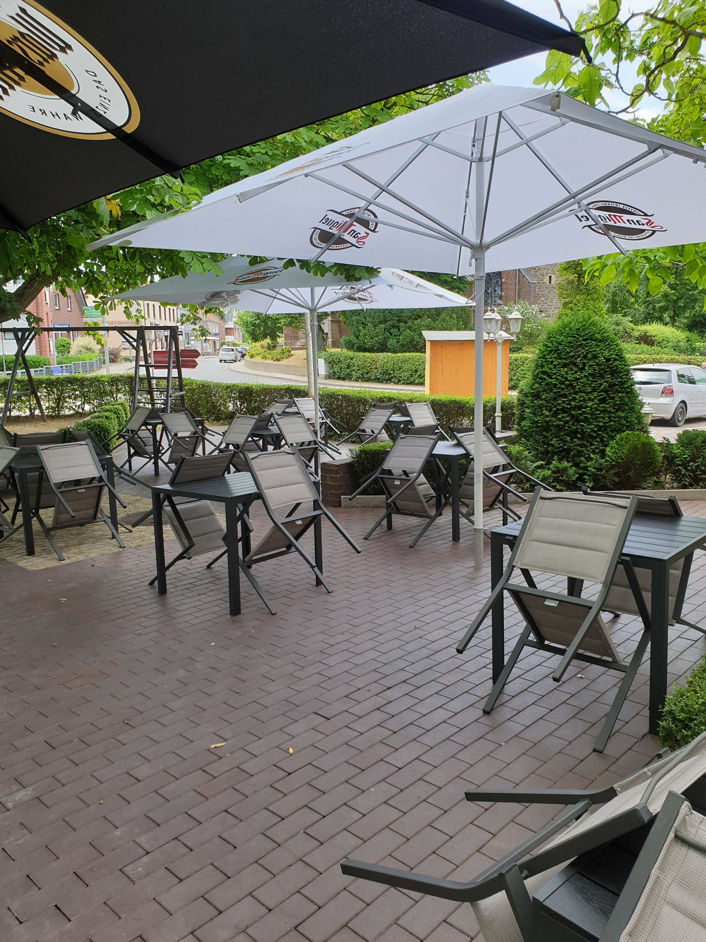 Un espace extérieur aménagé avec des tables et des chaises sous des parasols, un sol pavé de briques et une vue sur la rue et la verdure en arrière-plan.
