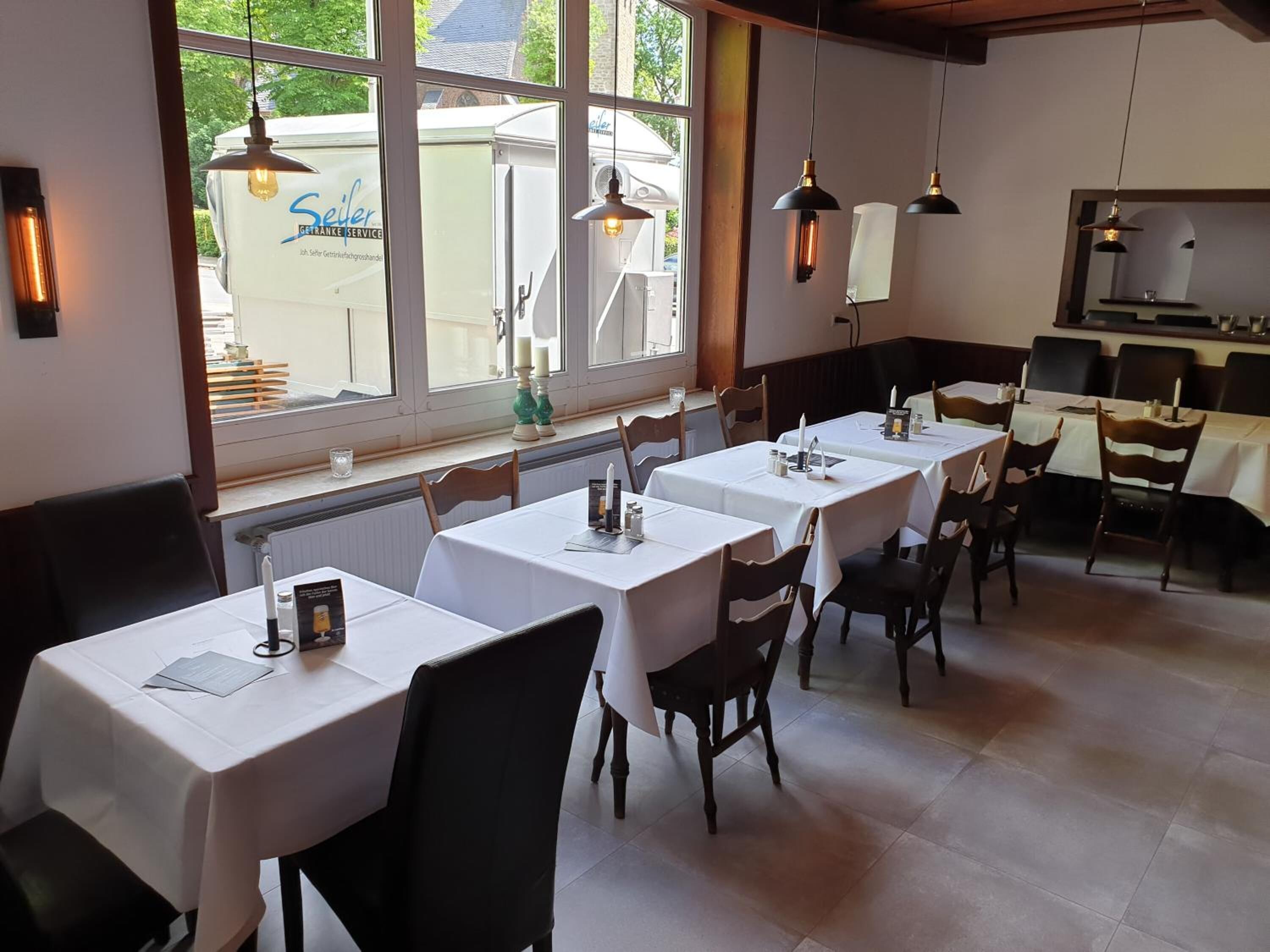 Ein Restaurant mit mehreren Tischen und Stühlen, großen Fenstern mit Blick nach draußen, Pendelleuchten und einem durch die Fenster einsehbaren Küchenbereich.