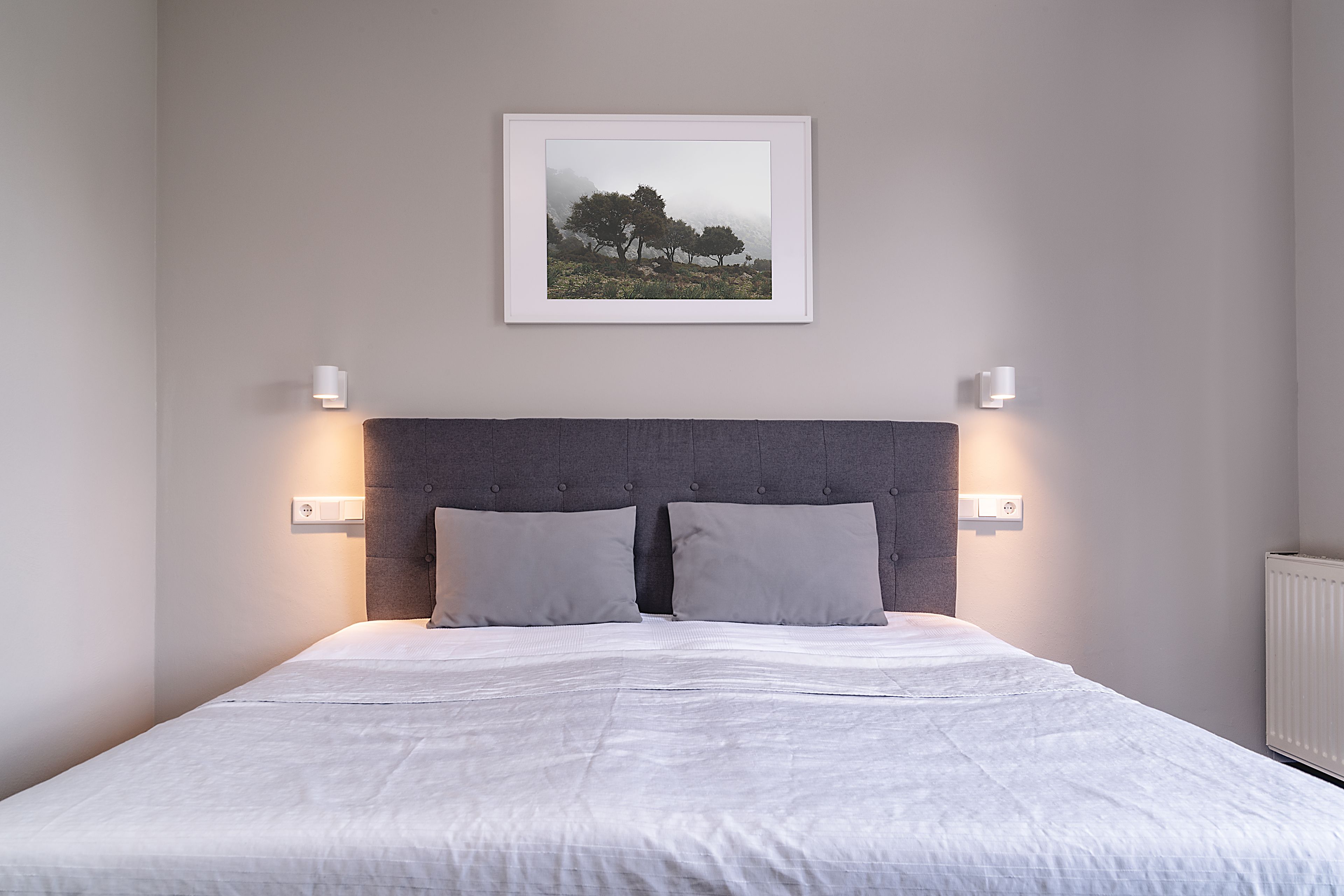 Quarto com cama, cabeceira, duas luzes fixadas na parede e um quadro enquadrado.