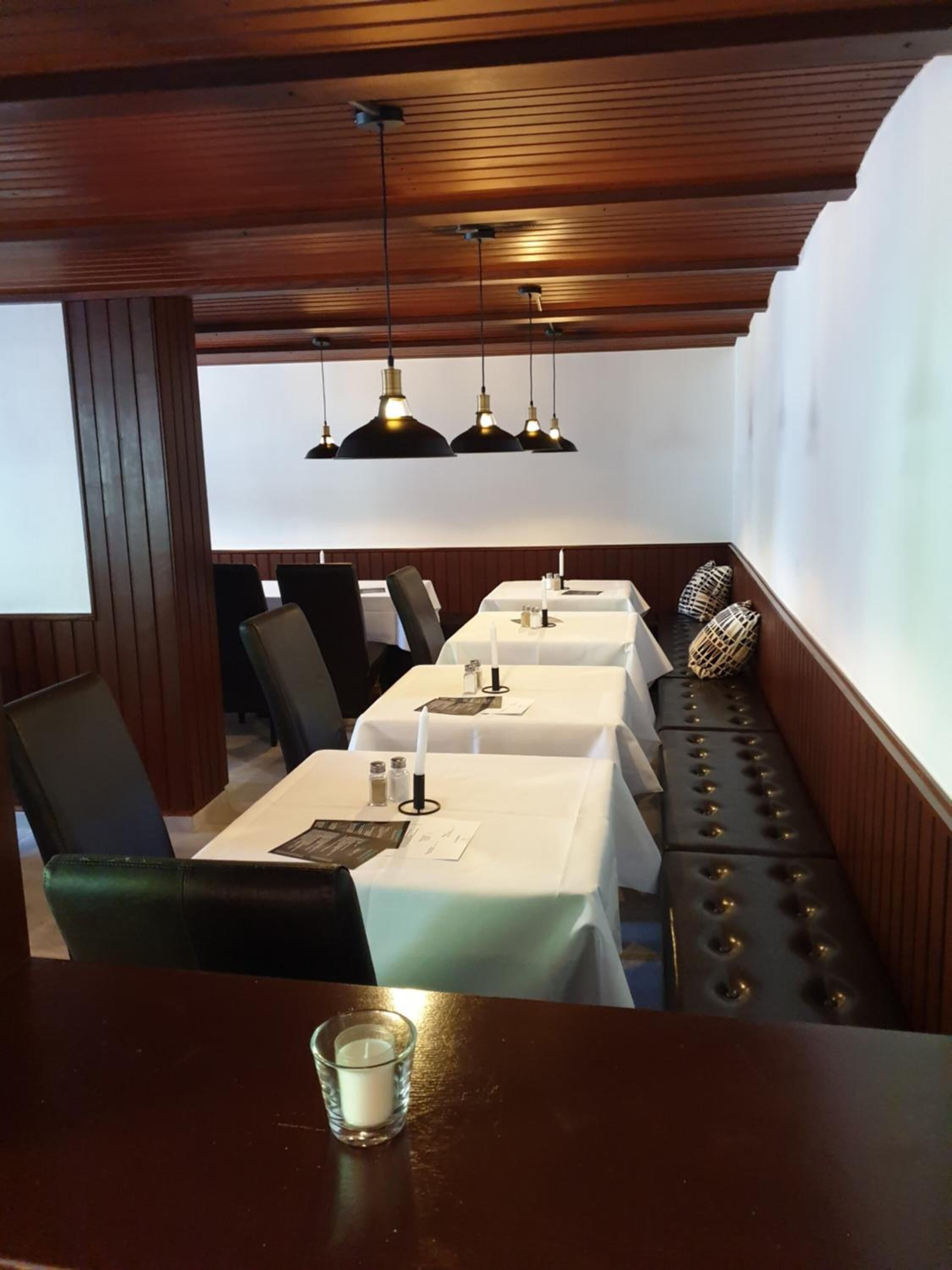 A restaurant with wooden ceiling beams, black upholstered chairs, white tablecloth-covered tables, and black pendant lighting.