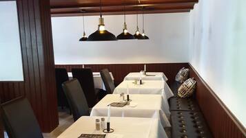 A restaurant with wooden ceiling beams, black upholstered chairs, white tablecloth-covered tables, and black pendant lighting.