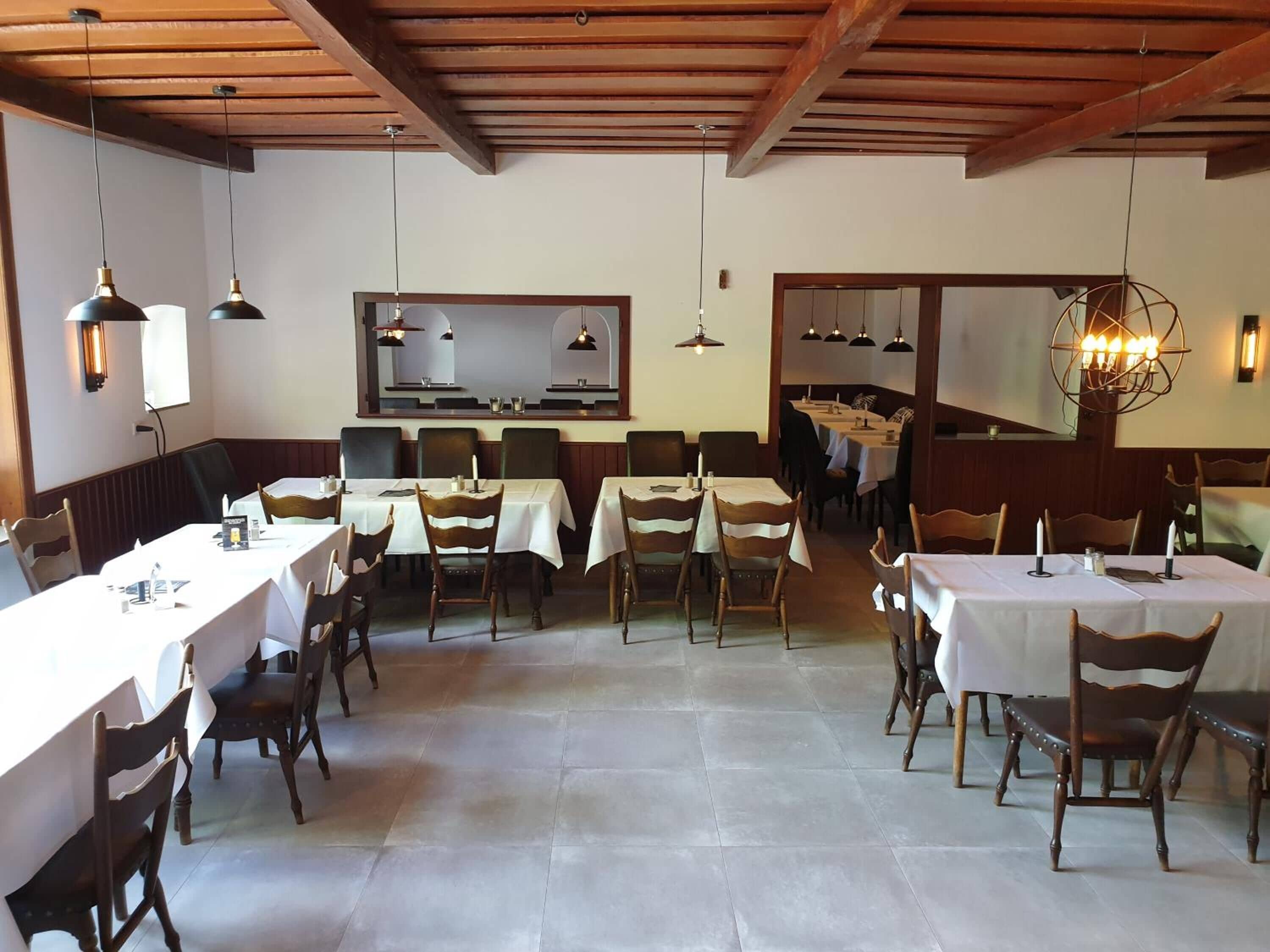 A restaurant with multiple tables and chairs, wooden beams on the ceiling, and pendant lighting.