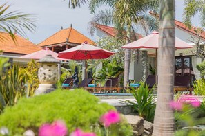 Zona junto a la piscina con sillones de descanso y sombrillas, rodeada de palmeras y plantas tropicales.