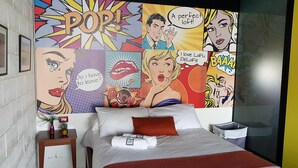 Een hotelkamer met een grote muurschildering in stripboekstijl, een bed met witte lakens en een rode kussen, een klein nachtkastje met een vaas, en een nachtkastje met een lamp.