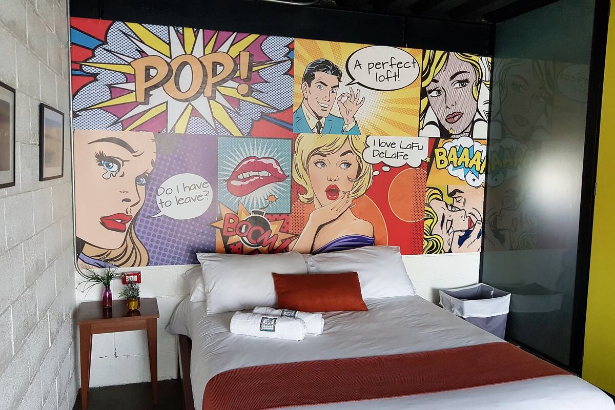 A hotel room with a large comic-style mural on the wall, a bed with white linens and a red accent pillow, a small bedside table with a vase, and a nightstand with a lamp.