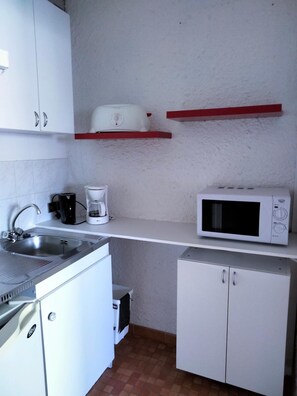 Une cuisine compacte avec des meubles blancs, un évier en acier inoxydable, une machine à café, un four à micro-ondes et une étagère rouge.