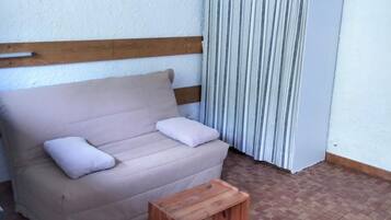 A room with a beige sofa, two pillows, a wooden side table, a striped curtain, and a framed picture on the wall.