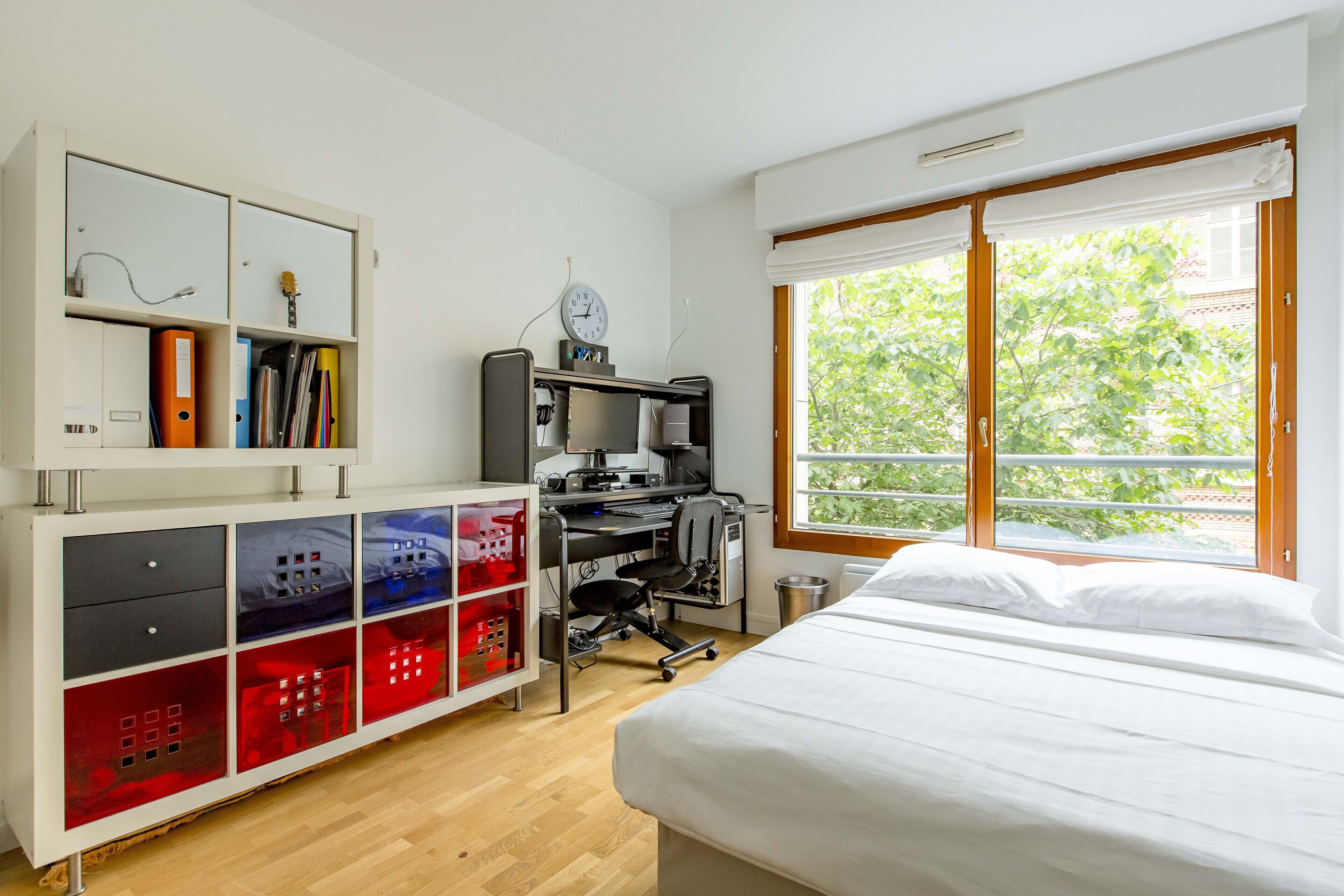 A bedroom with a bed, a desk with a computer, a bookshelf, and a window with a view of trees.