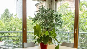 Et spiseområde med et træbord og fire stole, en potteplante samt store vinduer med udsigt til grønne områder og bygninger.