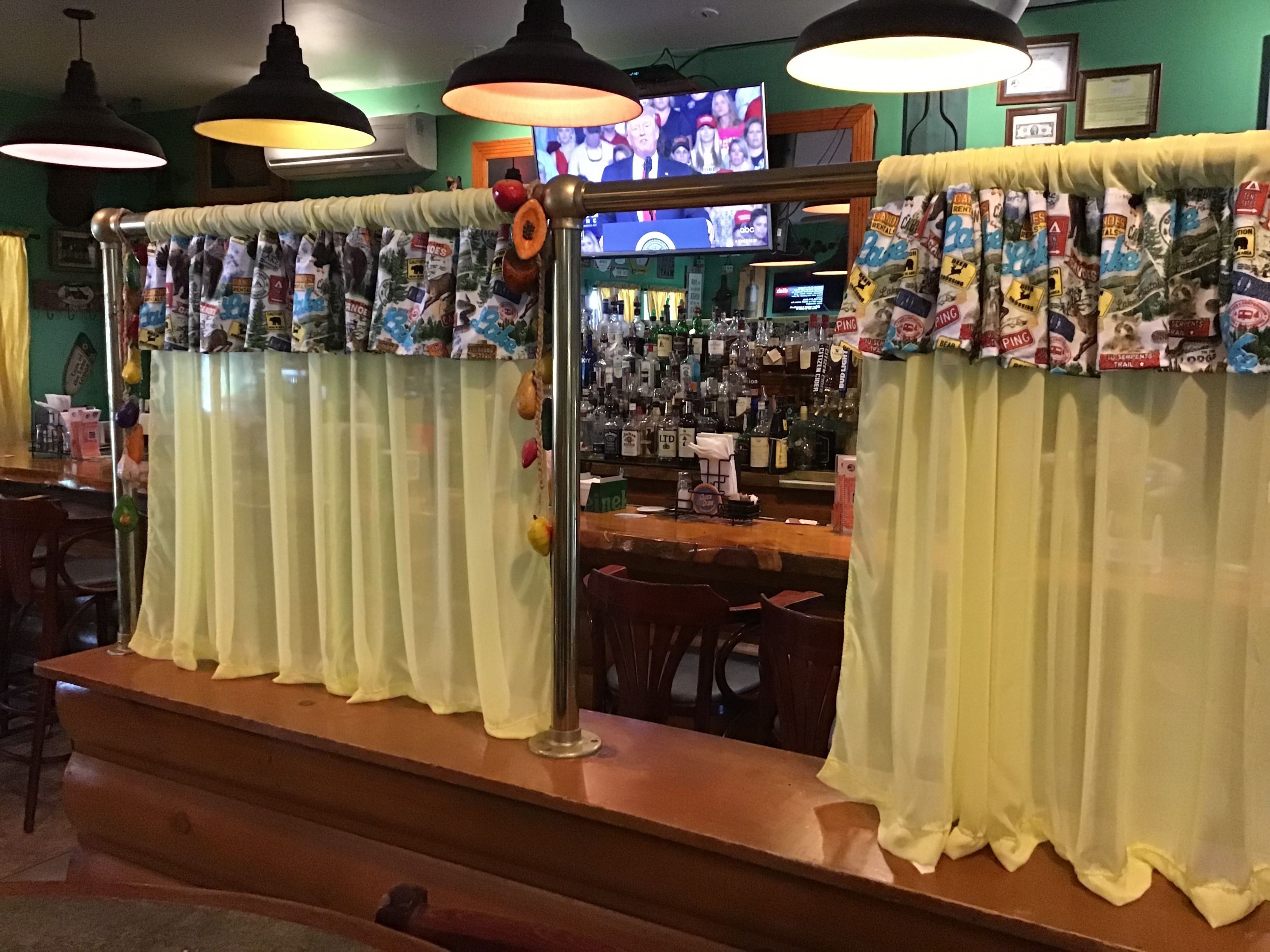 A bar area with yellow curtains, a television mounted on the wall, and a bar stocked with various bottles.