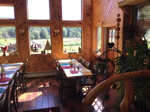 A wooden cabin with a dining area featuring a long table and chairs, large windows with a view of the outdoors, and a rustic decor including a wooden chair and a plant. - The Halfway House Motel (Elizabethtown)