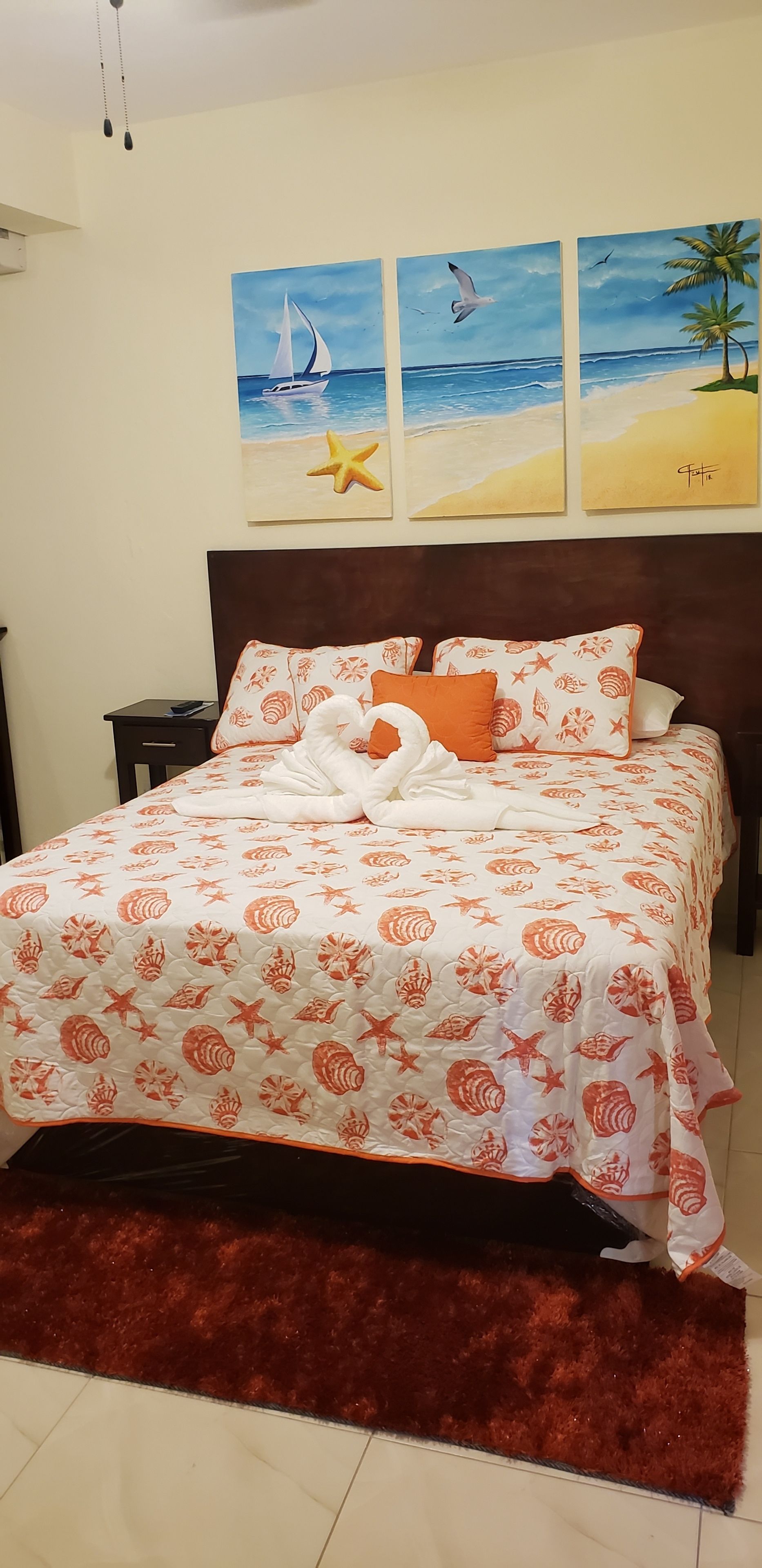 A bedroom with a bed featuring a seashell pattern bedspread, a wooden headboard, and a red rug. There are three paintings on the wall depicting beach scenes.