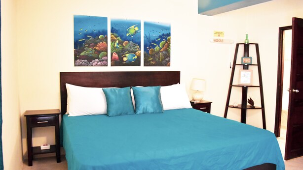 Een bed met een blauwgroene sprei en witte kussens, een nachtkastje met een lamp, en drie lijstjes met afbeeldingen van onderwaterlandschappen aan de muur.