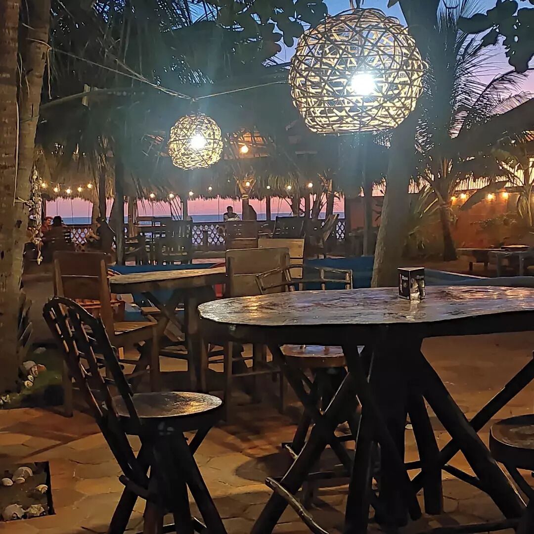 An outdoor seating area with round tables and chairs, thatched umbrellas, and hanging light fixtures.
