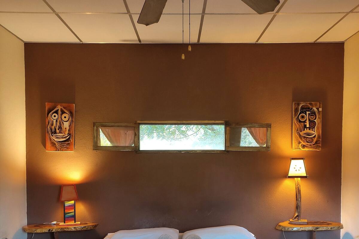 Ein Hotelzimmer mit einem Bett, einem Deckenventilator, zwei Nachttischen mit Lampen und Bildern an den Wänden.