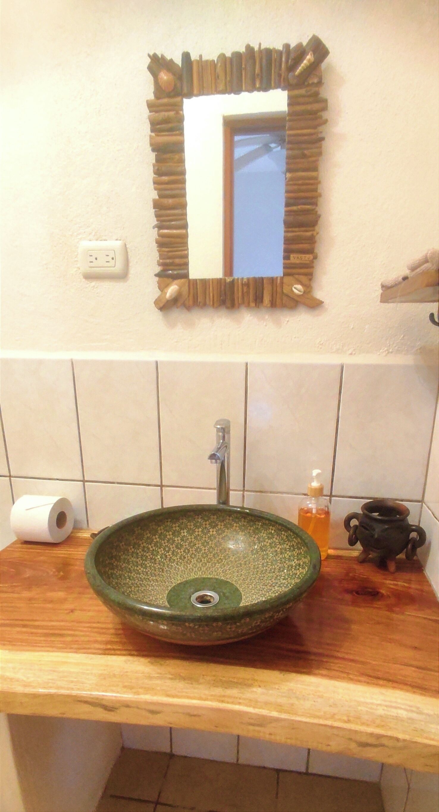 A bathroom with a wooden vanity, a green ceramic sink, a mirror with a rustic frame, a toilet paper roll, a soap dispenser, and a small black pot.