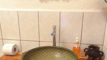 A bathroom with a wooden vanity, a green ceramic sink, a mirror with a rustic frame, a toilet paper roll, a soap dispenser, and a small black pot.