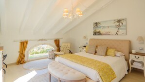 A bedroom with a large bed, a bench, a chair, a nightstand, a lamp, and a painting of a beach scene.