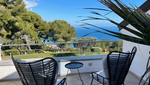 A balcony with two black wire chairs and a small round table, overlooking a scenic view of trees and the sea.