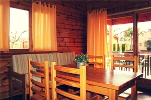 Interior de una cabaña de madera con una mesa de comedor, sillas, un banco y una ventana con cortinas.