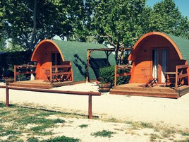 Dos cabañas de madera con techos verdes, cada una con un porche amueblado con muebles de madera y plantas en macetas.