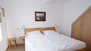 Een netjes opgemaakt bed met witte lakens in een kamer met een houten hoofdbord, een klein houten krukje en een houten kledingkast.