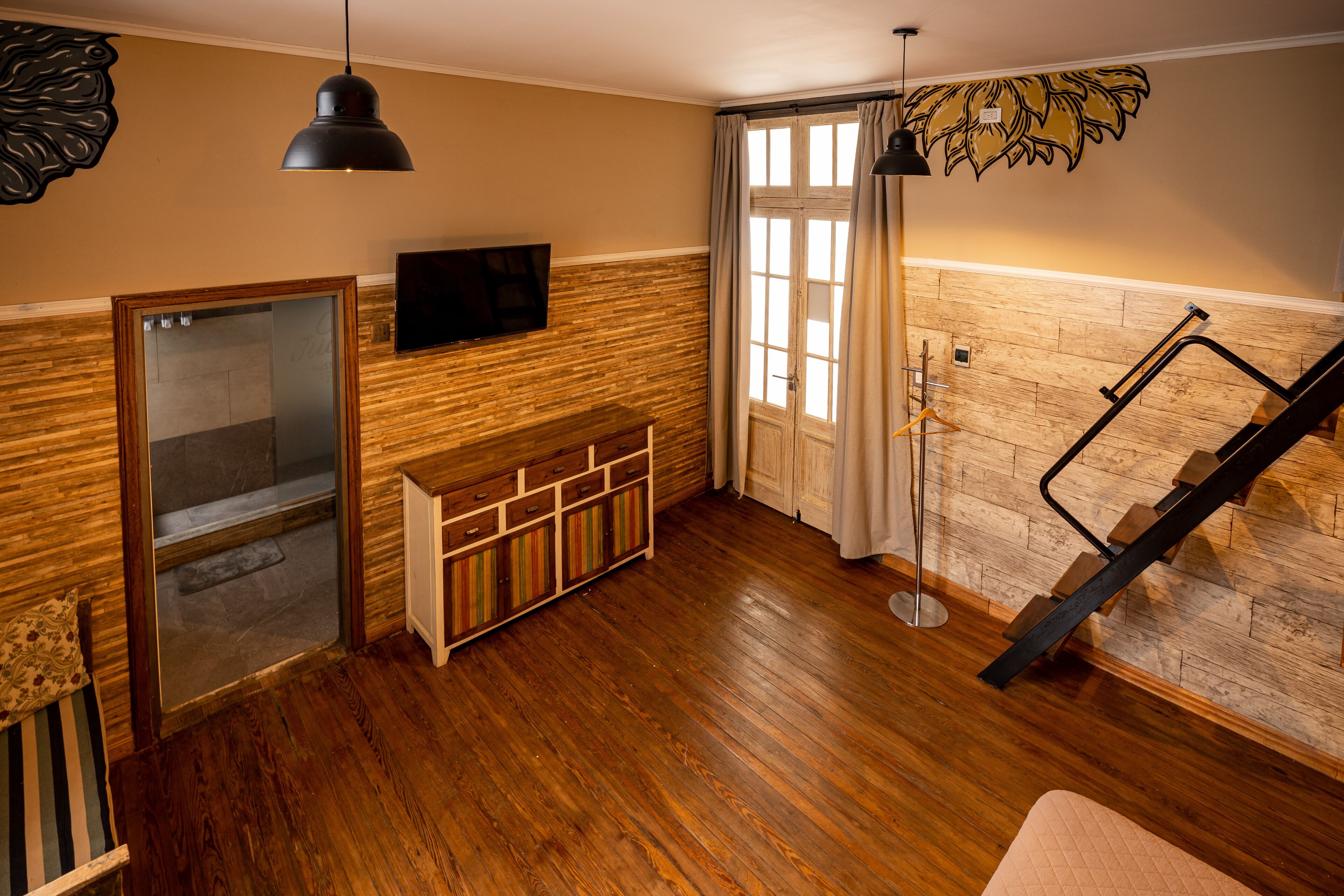 Una habitación con piso de madera, un banco de madera, un televisor montado en la pared, una escalera con barandilla metálica y una ventana con cortinas.