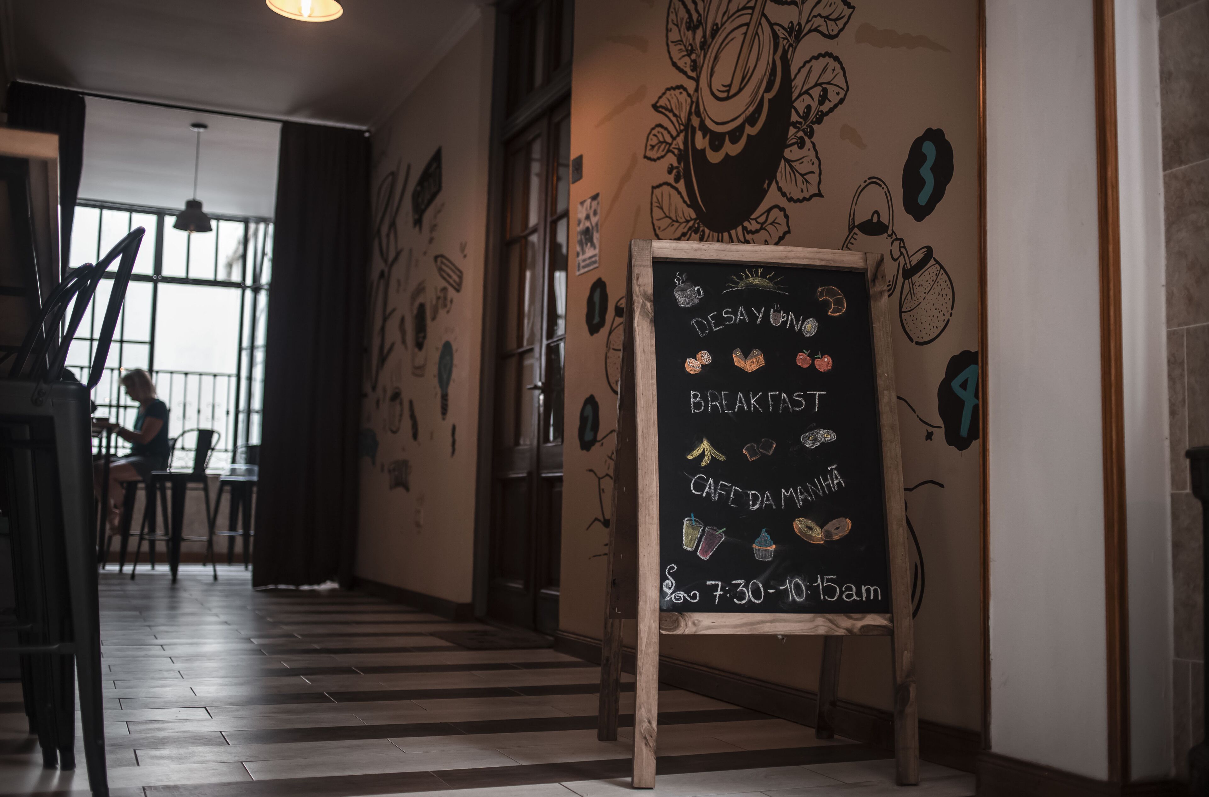 Una cafetería con un pizarrón anunciando los horarios de desayuno y 'cafe da manhã', suelos de madera y un mural en la pared.