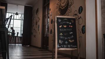 Una cafetería con un pizarrón anunciando los horarios de desayuno y 'cafe da manhã', suelos de madera y un mural en la pared.