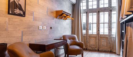 A room with a wooden wall, a desk, two chairs, a large window with curtains, and a framed picture on the wall.